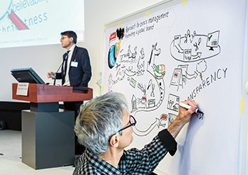 Conference at the Centre for Global Dialogue – One man drawing on a board while another one is speaking in the background (photo)